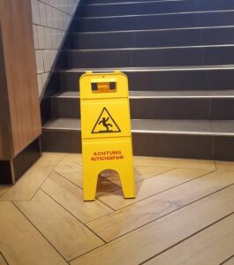 Ein gelbes "Vorsicht nass" Schild auf einem Holzboden symbolisiert Arbeitsschutz.
